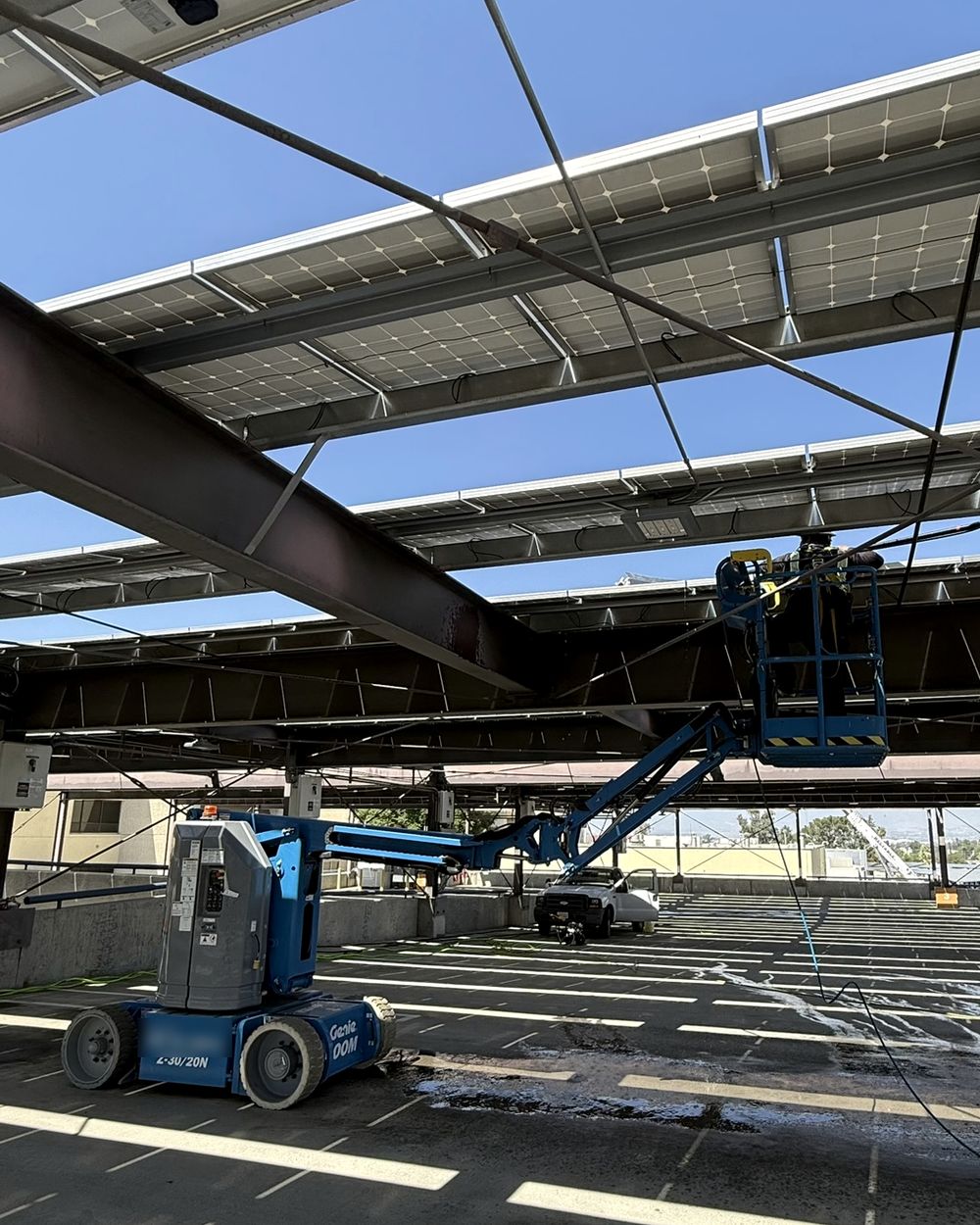 Technician in a boom lift cleaning a solar canopy over the top deck of a parking structure